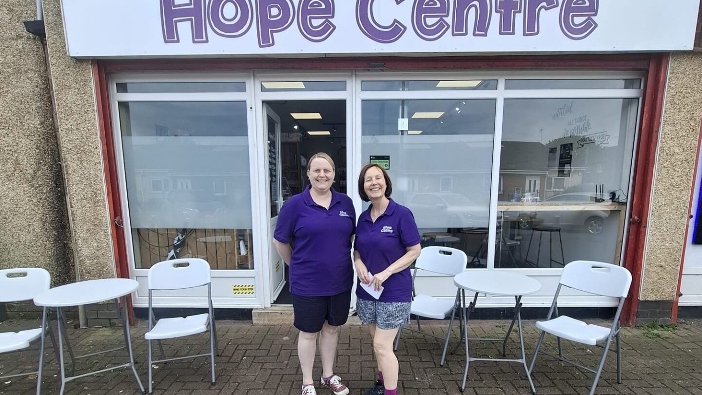 Two members of staff from the Hope Centre community food pantry dressed in purple polo shirts standing outside the front of the Hope Centre shop