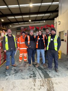 Image shows a group of 6 people, most of which are wearing Antlers standing in the FareShare Greater Manchester Warehouse and smiling at the camera.