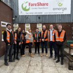 A group photo of 8 corporate volunteers, all wearing orange hi vis vests standing outside FareShare Greater Manchester's HQ under the organisation's sign