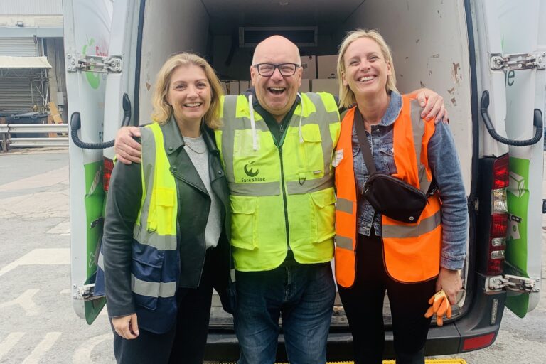 Image shows three people standing at the back of a FareShare Van. In the centre is a FareShare Greater Manchester driver standing with his arms around the shoulders of two women who are N Brown colleagues who have just finished making deliveries on a corporate volunteering day. All three are smiling at the camera