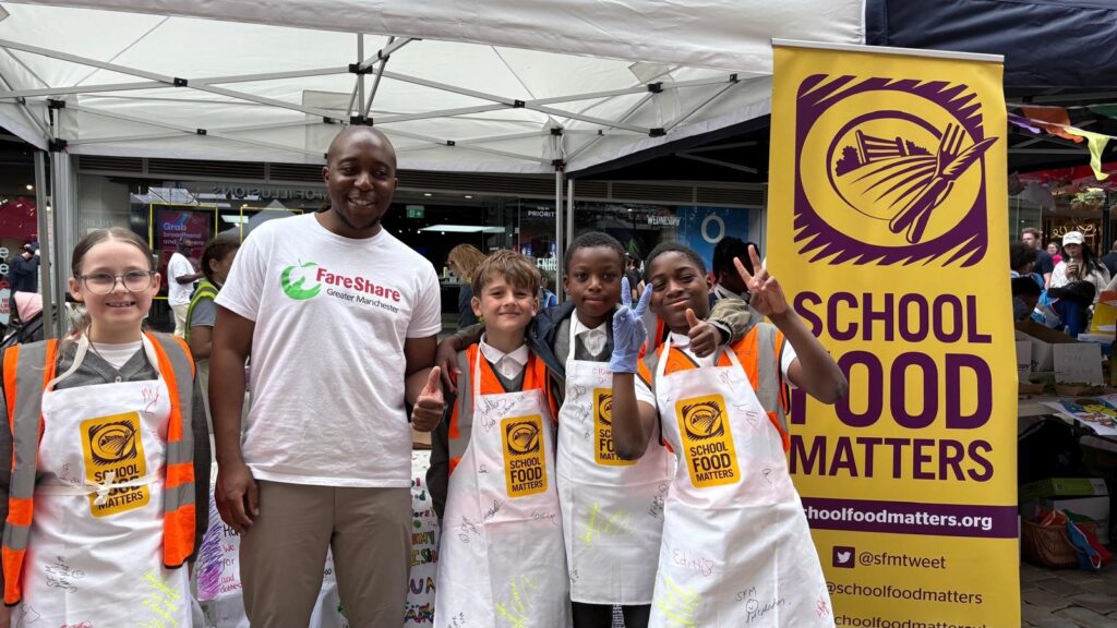 Image shows Kuda from FareShare Greater Manchester along with some of the Young Marketeers standing in front of their stall and smiling to the camera