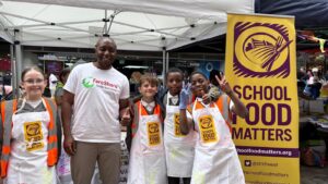 Image shows Kuda from FareShare Greater Manchester along with some of the Young Marketeers standing in front of their stall and smiling to the camera