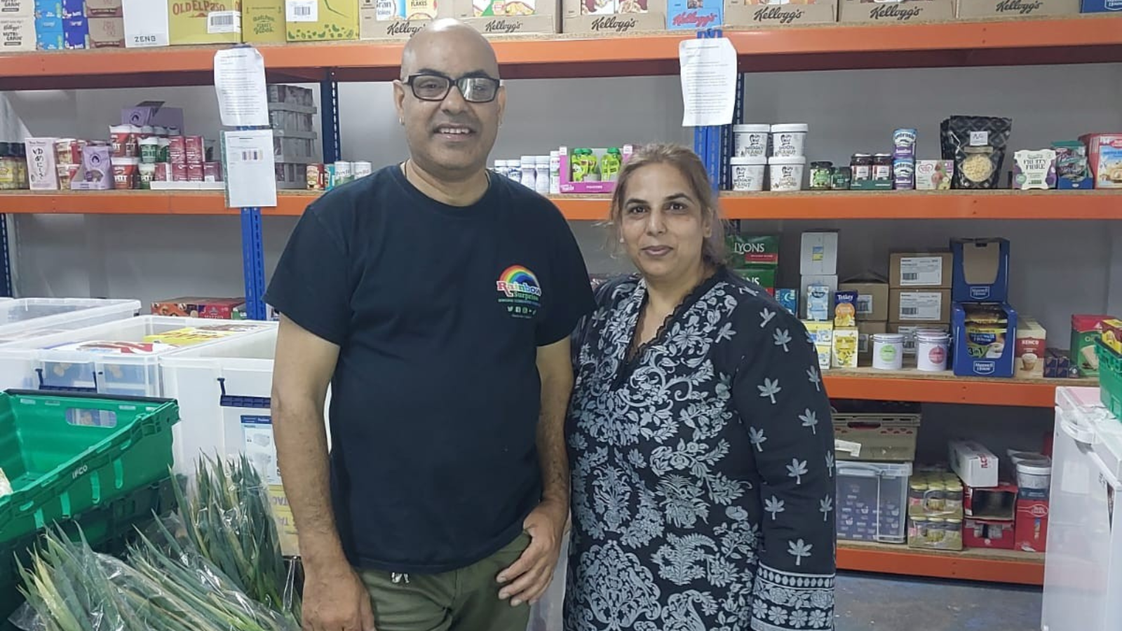 Image shows Shakar and Shabnam from Rainbow Surprise standing together and smiling at the camera in a large warehouse style of room filled with food on shelving behind them