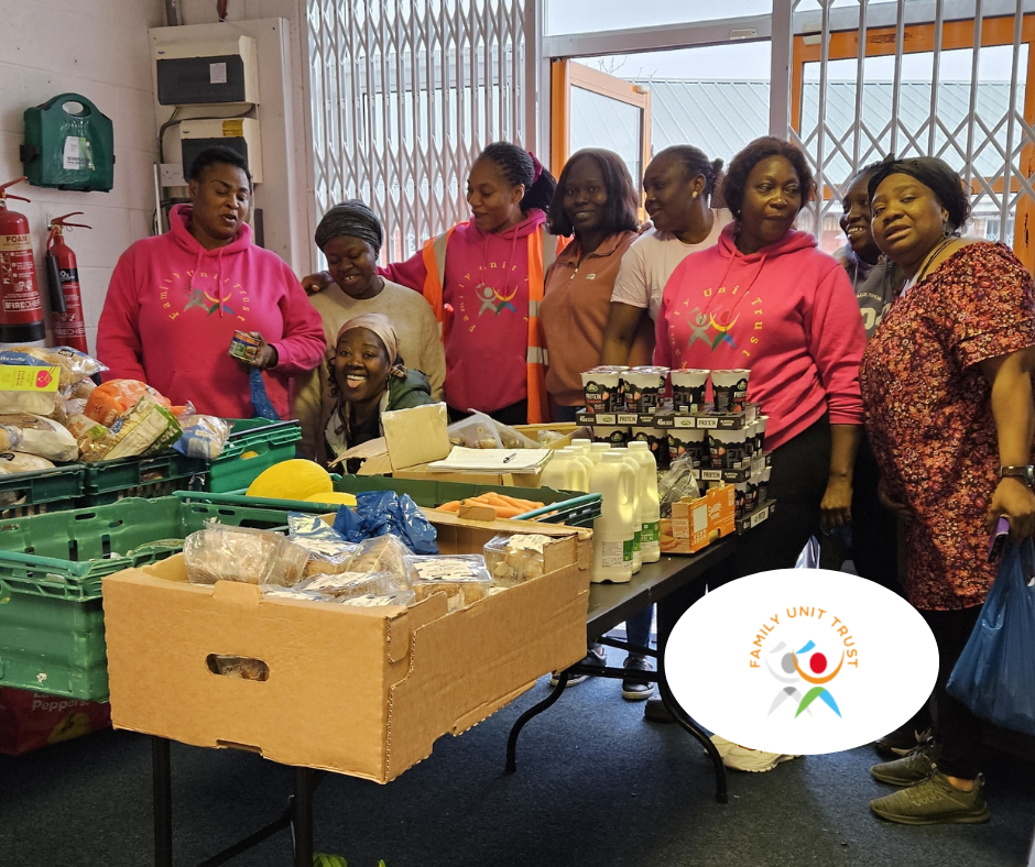 Image shows a group of women, some wearing pink sweatshirts with the Family Unit Trust Logo smiling and laughing behind a table covered with a food delivery